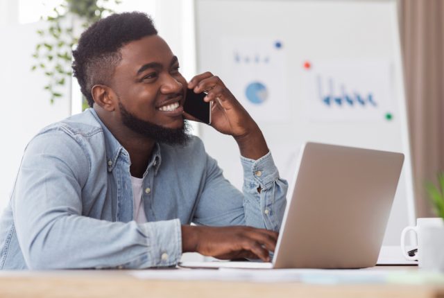 Cheerful man sat in front of his laptop and speaking on the phone