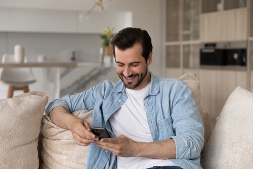 Man sat on couch in home, smiling when looking at mobile screen